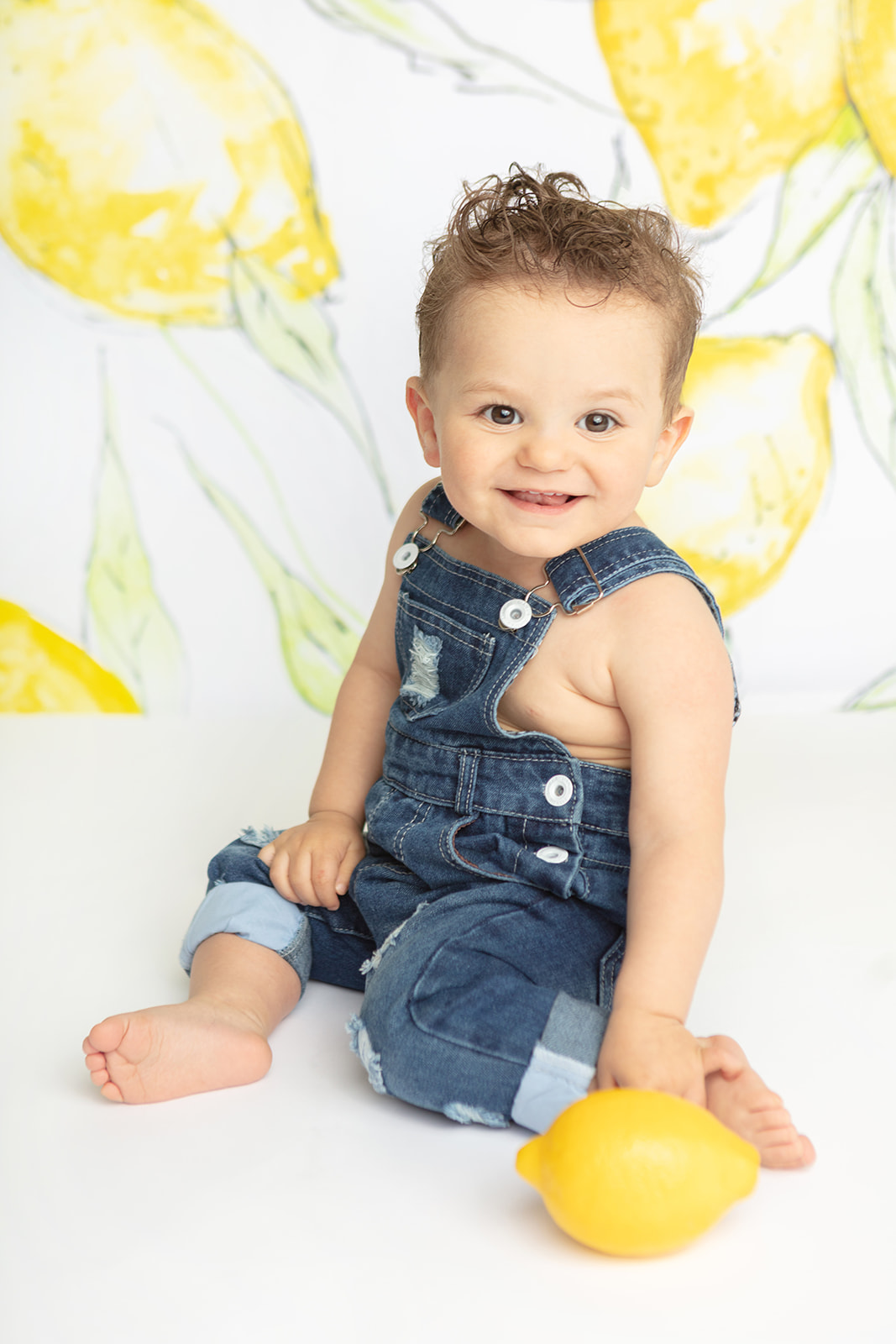 A smiling one year-old little boy in dark denim overalls leans slightly toward the camera. He has a mischievous smile on his face and is barefoot. A lemon prop sits in the foreground and the background is a painted lemon backdrop by Heidi Hope.