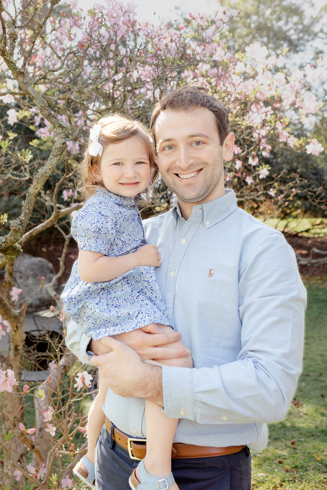 Connecticut dad wearing a classic chambray shirt, holding his little girl who is dressed in a ditsy floral print dress, her hair swept out of her face with a white grosgrain bow; behind the pair a tree blooms with delicate pink flowers