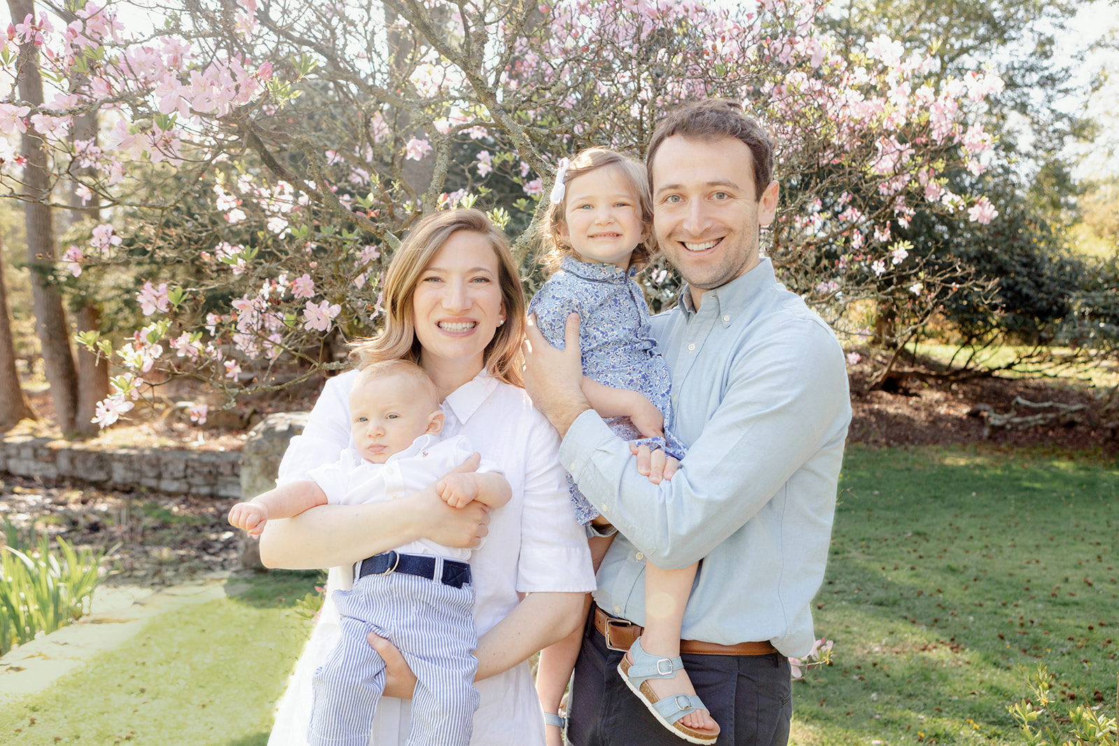 A Stamford, Connecticut-based physician couple hold their two children as mom, dad, and big sister smile at the camera. The family of four are photographed in a Connecticut park bursting forth with spring life.