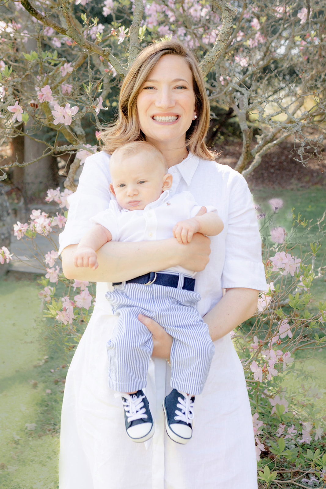 A New York physician mom, dressed in a classic white shirtdress, holds her infant son in front of her. The baby is also dressed in a crisp white Oxford, with seersucker pants, and the cutest navy Converse sneakers for babies. Behind the pair, a Connecticut tree brings forth delicate pink blooms.