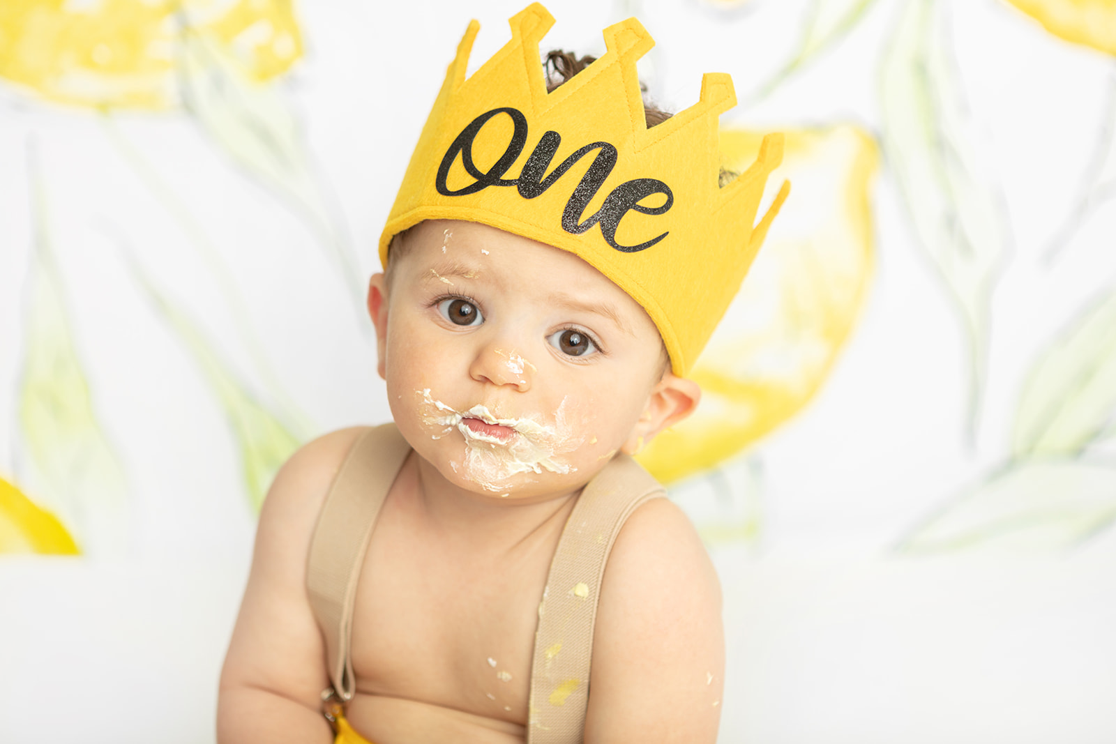 A one year-old little boy looks at the camera with a serious expression (with frosting and cake all over his face and suspenders). He wears a golden yellow first birthday crown. In the background is a handpainted lemon backdrop by Heidi Hope.