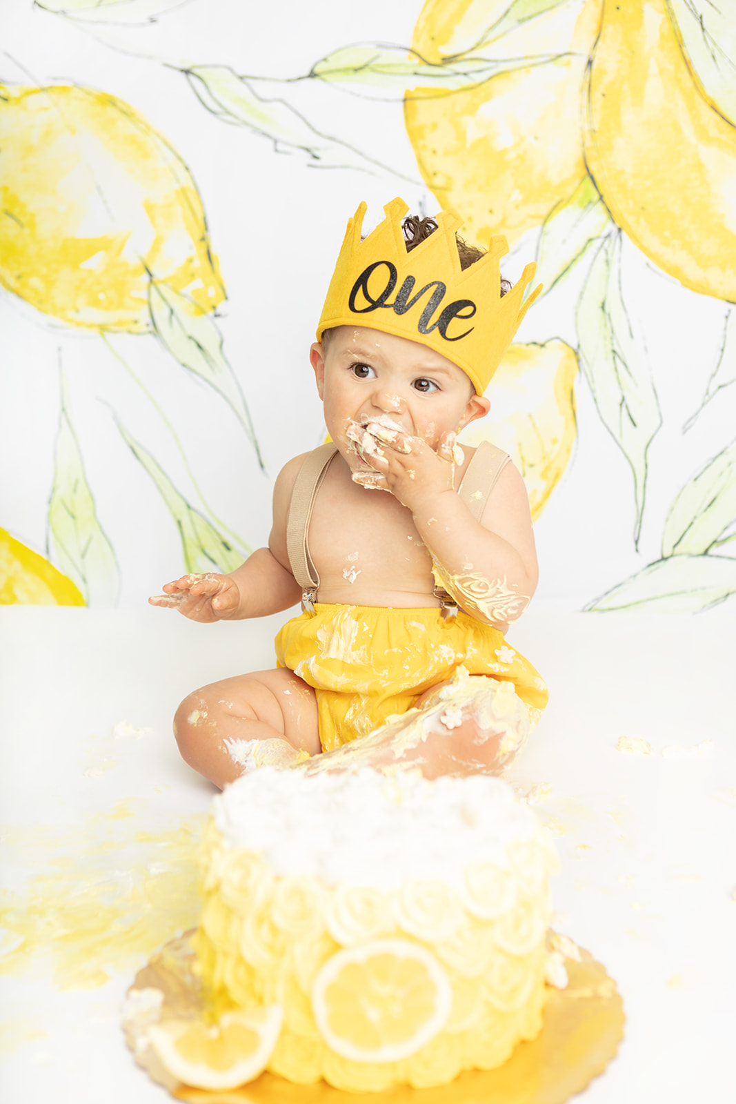 A one year-old little boy smashes cake into his mouth. He is absolutely covered in cake and icing! In the foreground of the picture sits a beautiful, ombre lemon smash cake. The backdrop is a handpainted lemon backdrop by Heidi Hope.