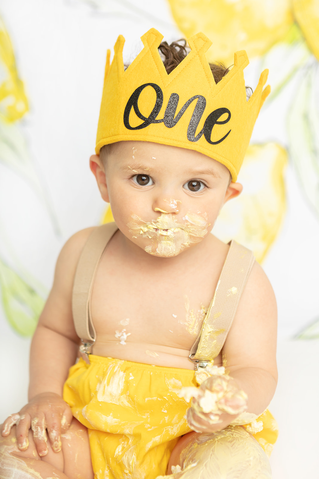 A little boy looks at the camera with a serious expression. He is wearing a golden yellow first birthday crown, suspenders, and yellow bloomers to match. He is absolutely covered in yellow lemon cake and frosting! In the background is a hand painted lemon backdrop by Heidi Hope.