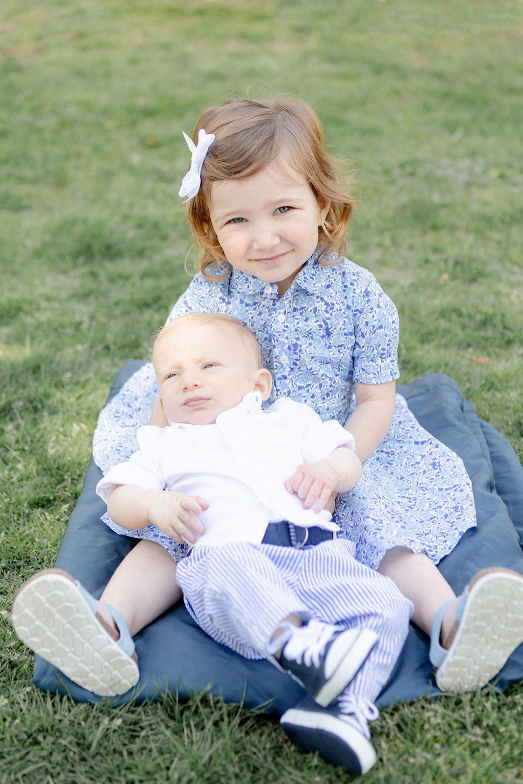 Lilah Sachs is photographed holding her baby brother in her lap. Her baby brother is quite squirmy, but the big sister calmly holds him and smiles at the camera, her cute sandals and feet naturally framing the image. The pair sit on a green, grassy field during springtime in Connecticut.