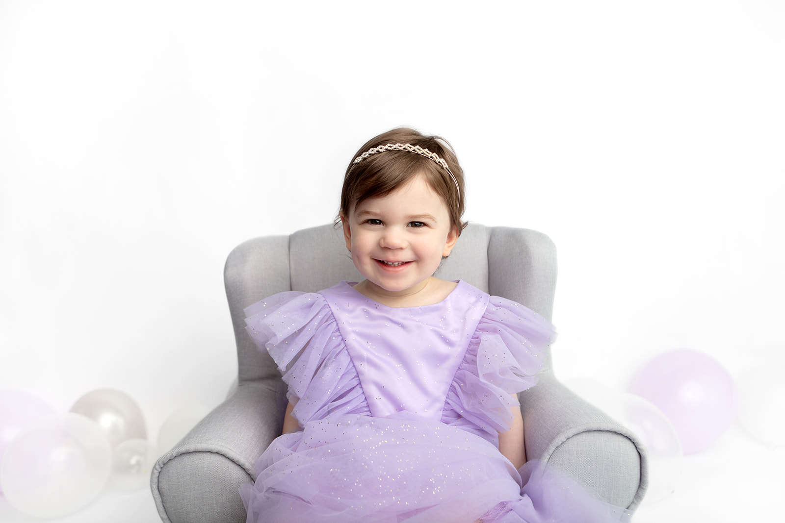 A two year-old girl celebrates her birthday with a milestone portrait session captured by Karen Kahn, of Looking Up Photography. The smiling girl wears a simple headband, and a lavender dress reminiscent of Molly Ringwald in Sixteen Candles. Lavender, silver, and translucent balloons pepper the white studio floor.