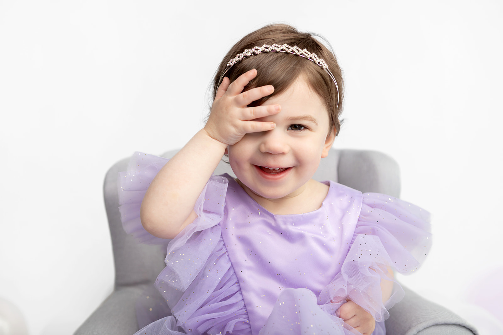 Two year-old Blake plays peekaboo with her photographer, Karen Kahn. Blake wears a bedazzled yet delicate headband, and a satin lavender party dress with tulle sleeves. Gold glitter sparkles in the foreground of Blake's two year portrait.