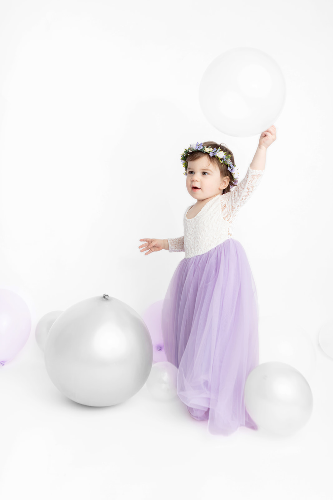 Two year-old Blake looks like a boho fairy princess in her ivory lace and lavender tulle gown. A white and purple flower crown adorns her head as she victoriously lifts a white baloon above her head. Silver, clear, and light purple balloons add depth to the photograph, captured at the Looking Up Photography studio in Greenwich, Connecticut.