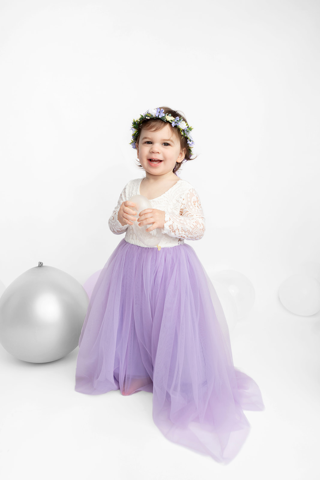 Two year-old Blake holds a small translucent party balloon and an easy smile on her face as she is photographed for her birthday. She wears a purple and white floral crown, and her wispy toddler hair peeks out from underneath it. Her cream lace and purple tulle fancy dress drapes on the floor of Karen Kahn's Greenwich, CT photography studio.