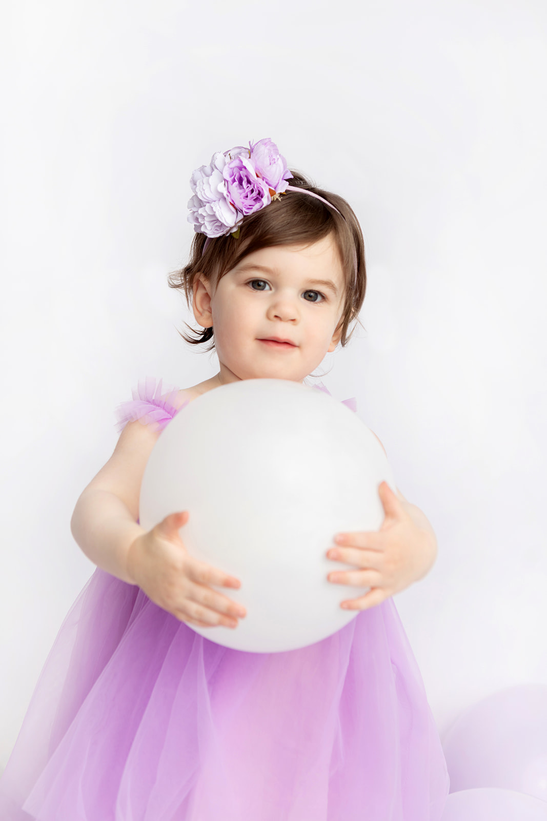 A two year-old little girl with short, chestnut brown hair, is photographed for her second birthday. She wears a delicate headband with a large purple flower on it, and holds a large white balloon in her arms like a playground ball. Her full, purple tulle party dress and lavender balloons bring visual interest to the modern milestone portrait.