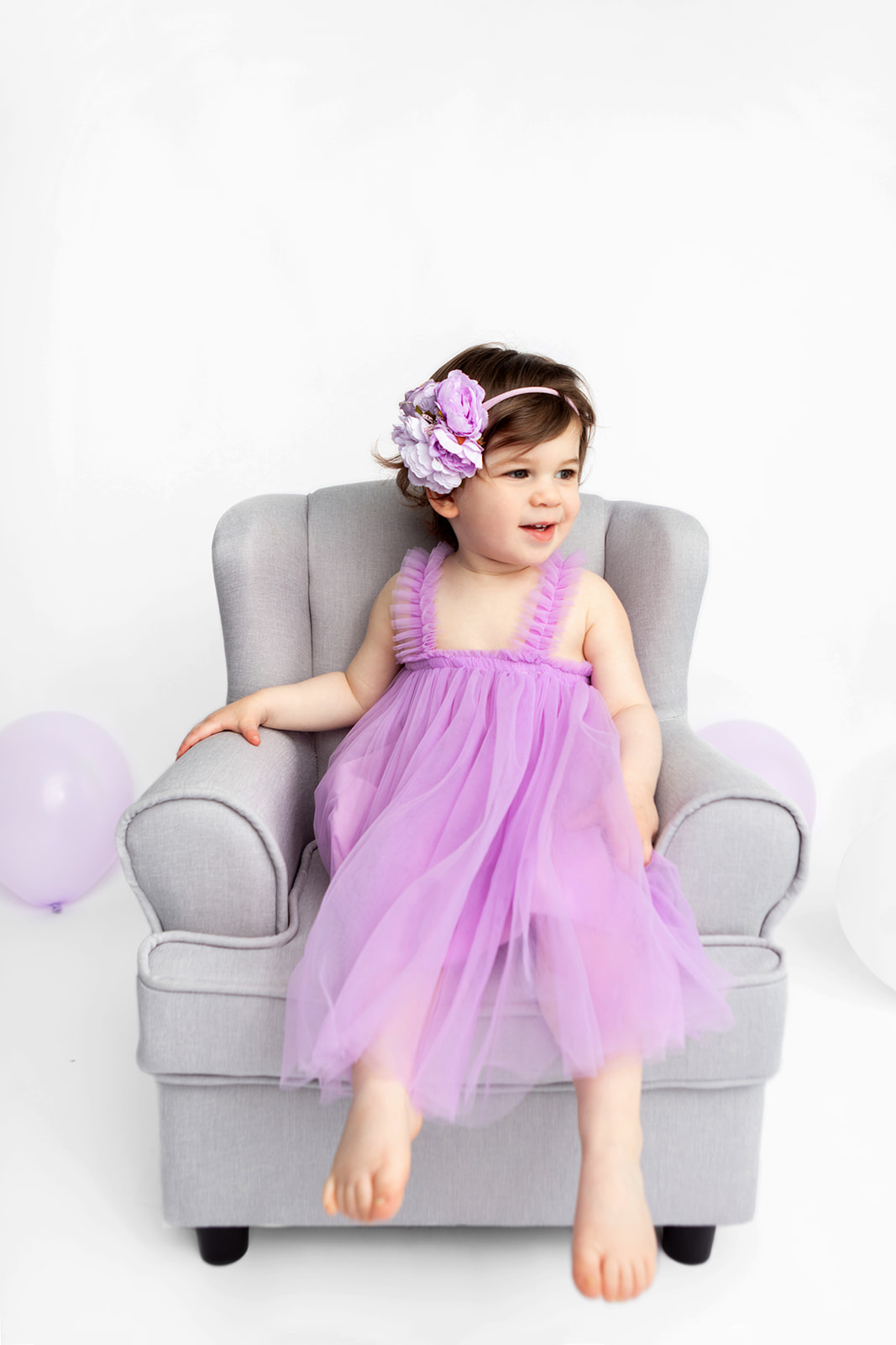 A cute little girl is photographed in motion, bare feet swinging from a gray tweed easy chair that's just her size. She smiles off-camera and her light purple party dress and large floral headband really pop, in this studio toddler milestone session photographed by Looking Up Photography, New York's Trusted Baby Photographer.