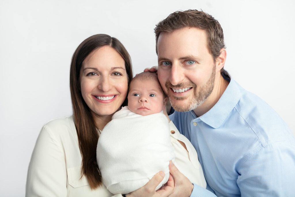 Proud parents hold up their newborn baby son in between them and smile. The newborn baby boy is swaddled in creamy white and has a serious expression on his face as he looks slightly off-camera