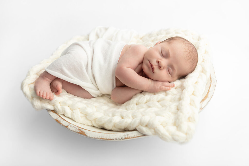 A newborn baby boy is lightly swaddled in cream as he lies on an ivory chunky knit blanket in a rustic washing bowl, against a soft white studio backdrop