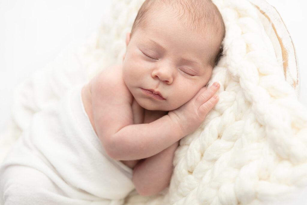 A newborn baby boy lies asleep with his cheek resting on his right hand, on an oversized cream colored crochet blanket. The baby boy is lightly swaddled in ivory and placed in a shabby chic bowl, photographed against a soft white studio background