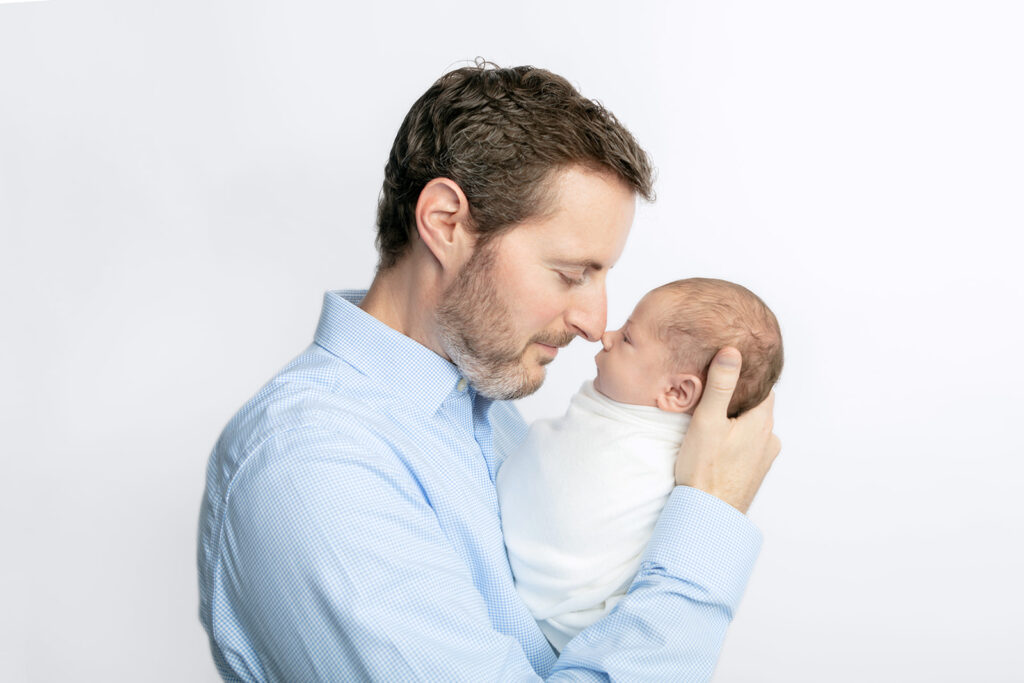 A dad with a short, salt and pepper beard, wearing a blue checkered shirt, holds up his swaddled newborn son to his face and the pair touch noses