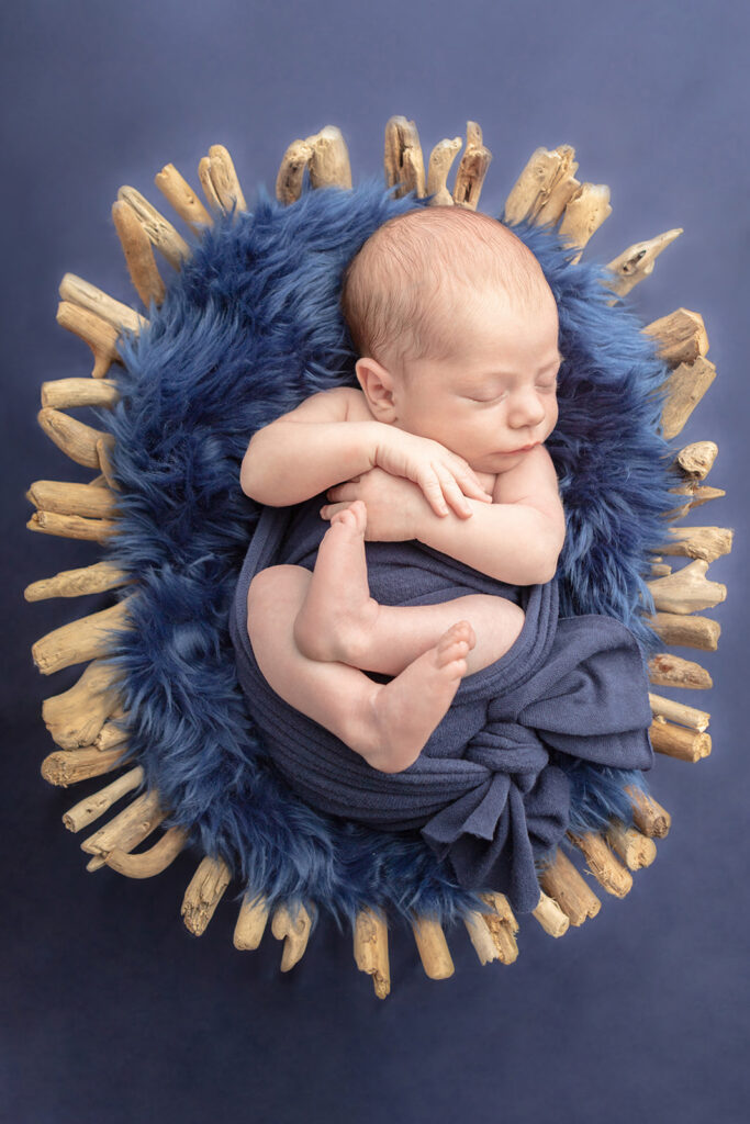 Newborn baby boy Landon lies sleeping in a classic newborn pose, swaddled with his arms and legs out, in a stretchy navy swaddle. He lies on a navy flokati in a driftwood basket, against a navy studio backdrop