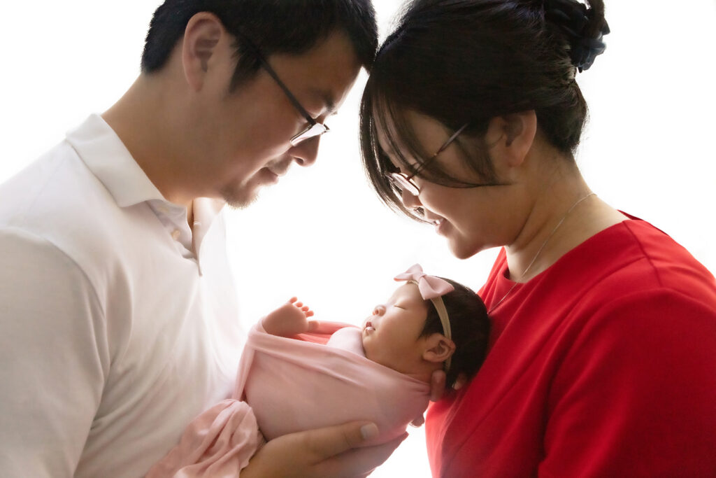 Proud mama and dada look peacefully down at their newborn daughter, who is being cradled in dad's hands. Her little newborn foot peeks out from the swaddle as she sleeps. Mama wears a red top while Dad wears a white polo. 
