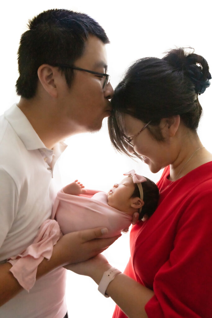 A proud dad and husband kisses his smiling wife on the forehead as she looks down at their newborn baby girl, swaddled in pink with a pink bow on her head.