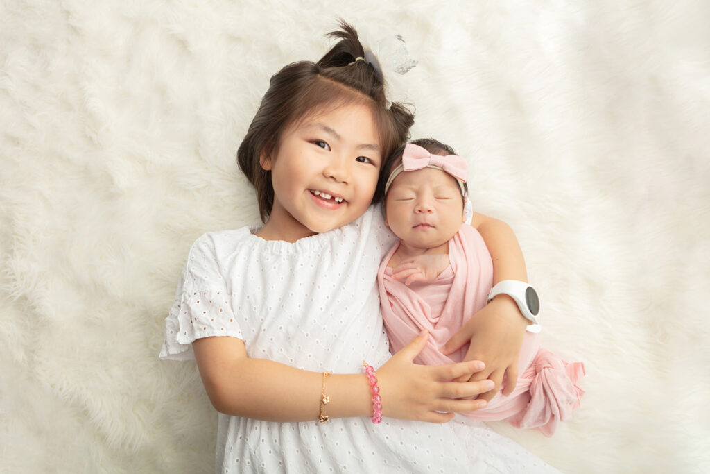 A proud big sister with a gap-toothed smile holds her newborn baby sister in her arms. The pair lie on a cream colored flokati rug. The big sister wears a white eyelet dress and little girls' jewelry, while her baby sister is lightly swaddled in light pink and wears a light pink bow.