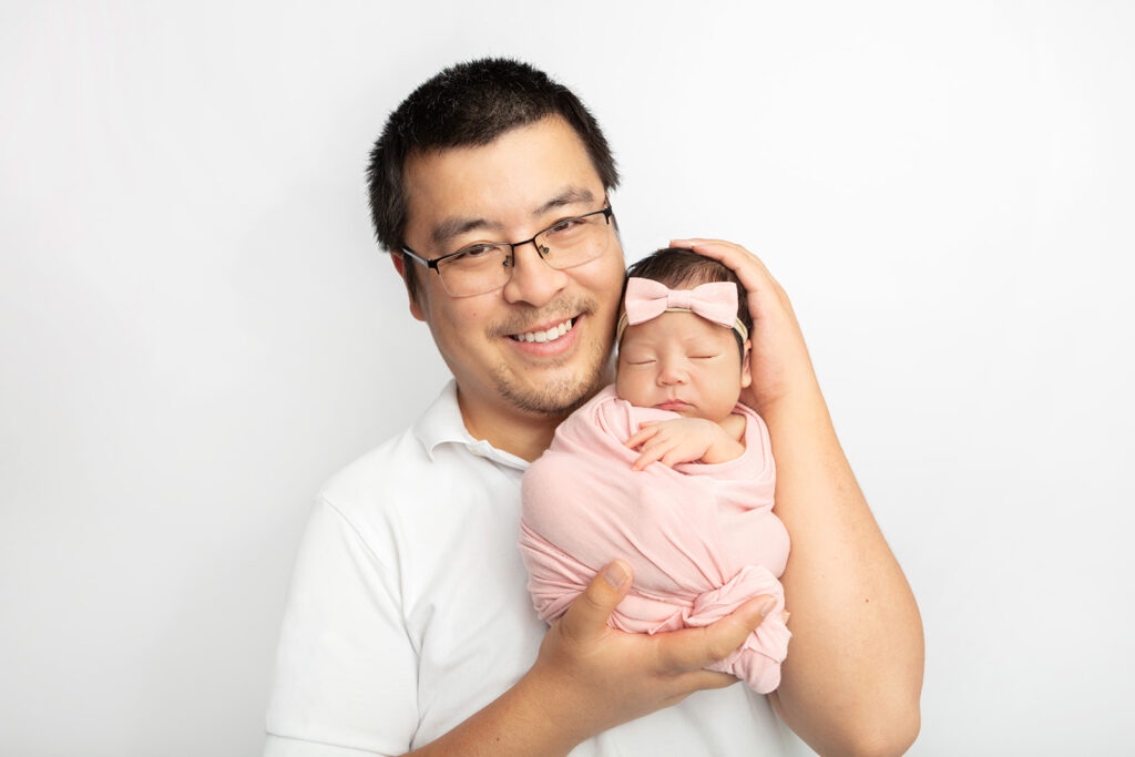 Proud father, COVID-19 scientist Dr. Nianshuang Wang, holds his newborn daughter up in his hands, with her head lightly resting on his cheek. Dr. Wang wears glasses and a white polo shirt, while his daughter is swaddled in pink and wears a pink bow.