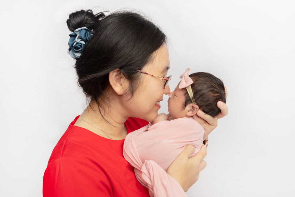 A mama dressed in traditional celebratory red holds her newborn baby girl up to her face and they touch noses. The mom is smiling, and wears tortoiseshell glasses and a silky blue scrunci in her hair.