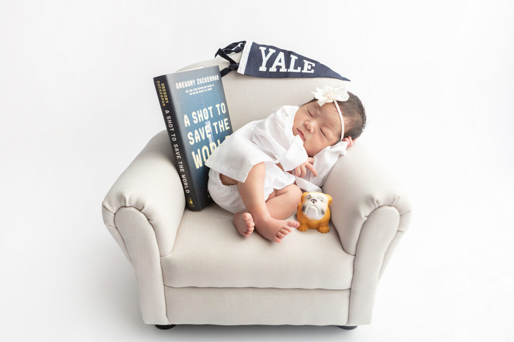 Dr. Nianshuang Wang family newborn session; a newborn baby girl sleeps on an infant sized overstuffed chair, with a Yale banner and bulldog, and the book A Shot to Save the World
