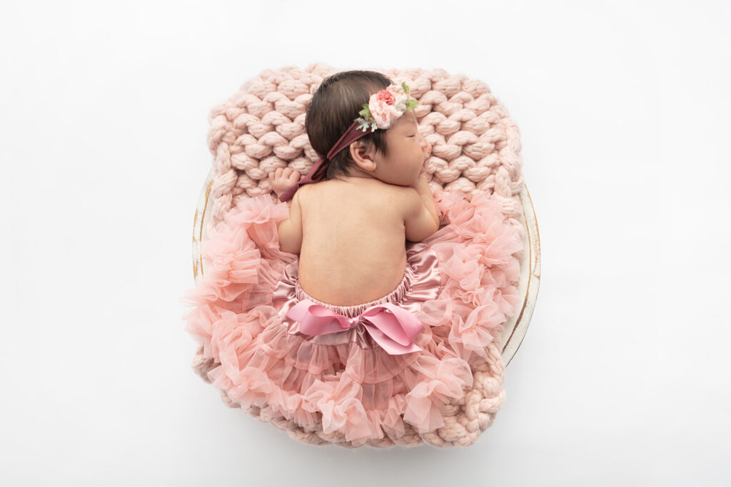 A newborn baby girl wearing a dusty pink tutu with a big bow on the back, lies on her right hand with her mouth slightly open, sleeping on a chunky knit, light peach colored blanket. The baby and blanket are in a shabby chic wash basin, photographed against a white backdrop.
