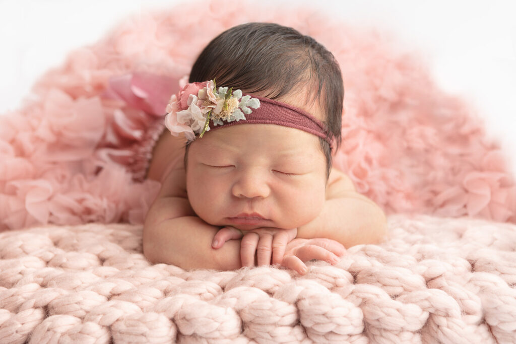 A newborn baby girl with chunky cheeks lies her head on her hands and folded arms as she sleeps on a big, chunky knit, light pink blanket. The baby girl has a head full of dark hair and wears a stretchy maroon headband, accented with dusty florals. Her tutu poofs out behind her, framing the image.