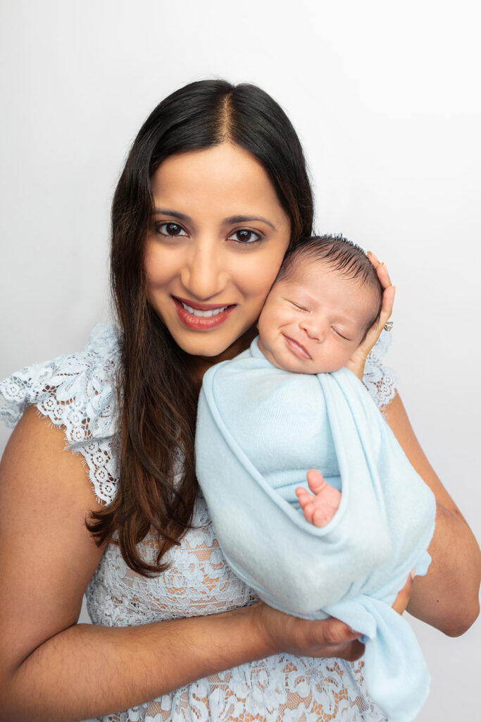 A beautiful multicultural mother with dark eyes and long, dark hair holds her smiling newborn baby boy up next to her face. The baby is swaddled in a felted, sky blue swaddle and Mom wears a matching eyelet dress.