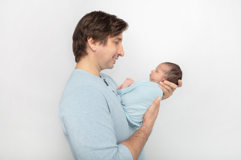 A father holds his newborn son in his hands and smiles down at him. The pair match: dad's henley shirt and the baby boy's swaddle are almost the same color of sky blue.