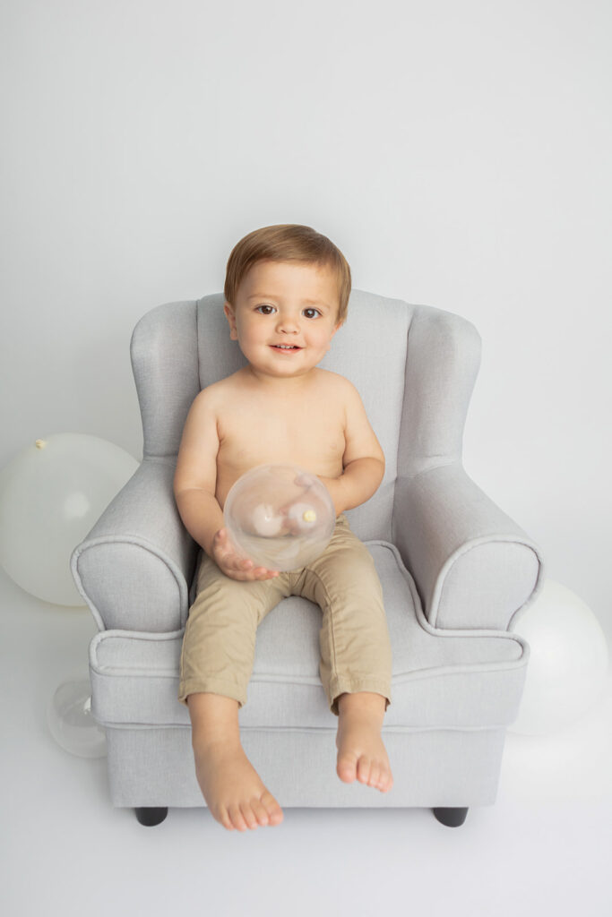A one year old boy with sandy colored hair sits in a light gray overstuffed chair that is just his size. He holds a translucent balloon in his hands and the white background also has a few balloons on the ground.