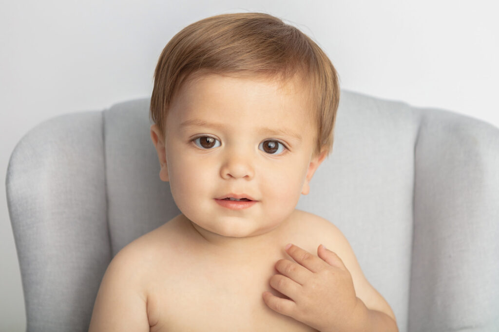 A one year old little boy with big brown eyes looks into the camera with a slight smile on his face, his little hand reaching up toward his chin. He is pictured in a light gray, comfortable chair sized for toddlers.