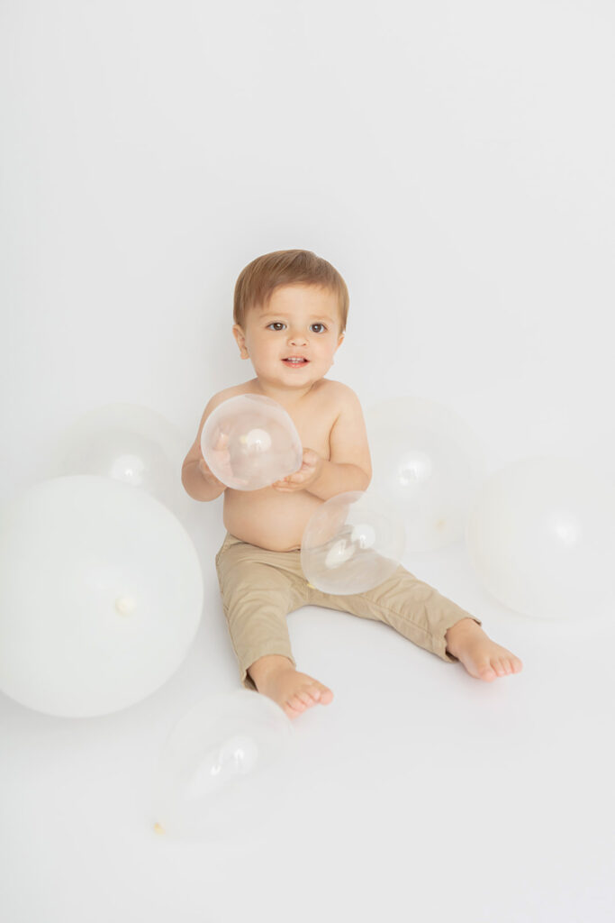 Baby boy Austin is photographed on his first birthday, sitting in a white studio setting, holding a clear balloon in his hands. Translucent and white balloons of different sizes cover the studio floor and Austin smiles, looking slightly off camera.