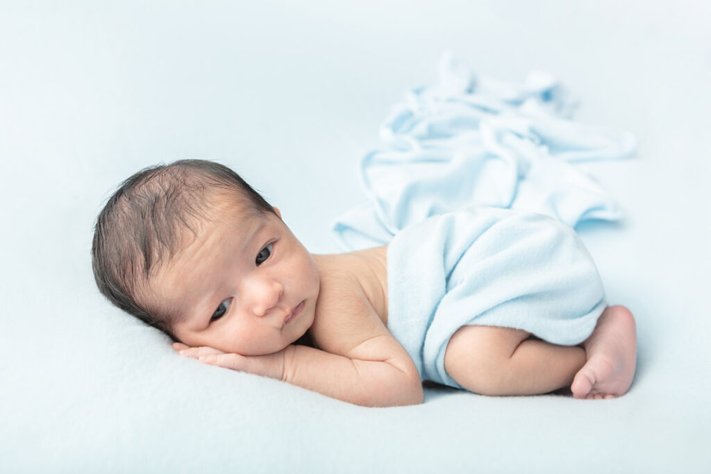 A newborn baby boy with dark hair rests his head on his hands. His eyes are open and he has a sleepy, almost bored expression on his face. His light blue swaddle trails behind him in an almost snowflake-like pattern.