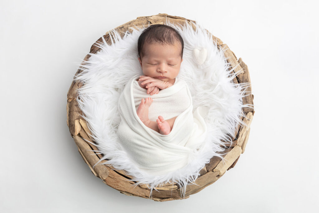 A newborn baby boy named Noah sleeps peacefully in a light swaddle. His hands and feet poke out of the swaddle in a relaxed way. The baby has a head full of dark hair.