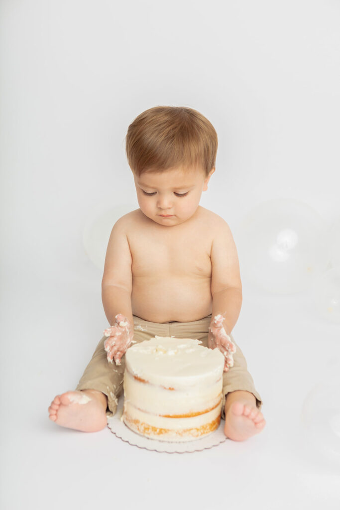 A one year old little boy looks down at his hands, which are covered in cake frosting, as if he's wondering what to do about the mess. In front of him sits a naked cake on the floor of the Looking Up Photography studio in Greenwich, Connecticut.