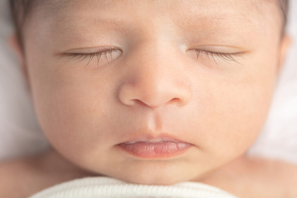 A macro style portrait of the face of a newborn baby boy named Noah. Baby Noah's chin rests lightly on a ribbed blanket. His facial features (including the longest eyelashes) are clear and soft-looking!