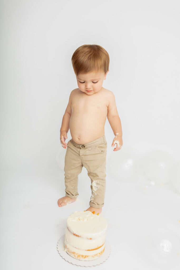 A one year old little boy with a slightly protruding belly looks down at his smash cake and smiles. He has icing on his hands, chino pants, and feet, and the floor of the studio is peppered with clear and soft white balloons.