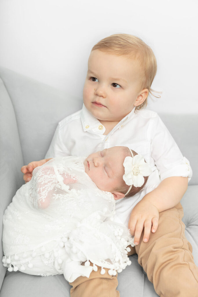 Big brother Alexander holds his newborn baby sister in his lap. He looks seriously off-camera. The pair sit on a light gray chair in the Looking Up Photography studio in Greenwich, Connecticut.
