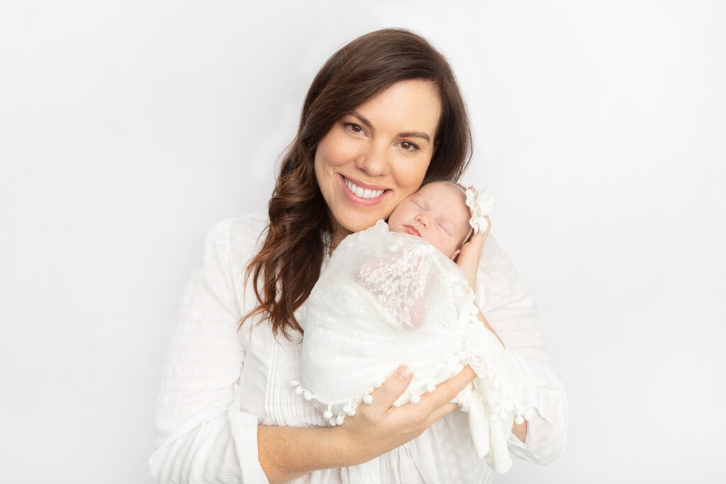 A beautiful new mom with dark eyes and hair smiles as she holds her sleeping newborn daughter up to her cheek. The baby girl sleeps comfortably, swaddled in lace.