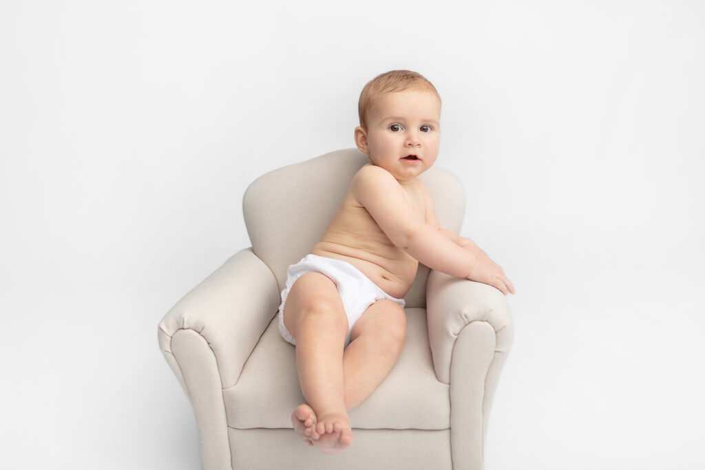 A Gerber-looking baby with short hair and big, dark eyes, looks expressively at the camera. The baby sits with ankles crossed in a baby sized overstuffed creamy leather chair against a white backdrop.