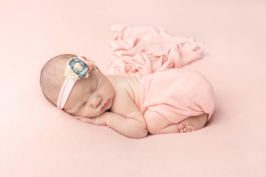 A newborn baby girl, Olivia, sleeps folded up like a little bean on a large, peachy pink pillow. She is lightly swaddled at the waist in a matching peachy pink, the swaddle blanket trailing behind her into the background of the image.