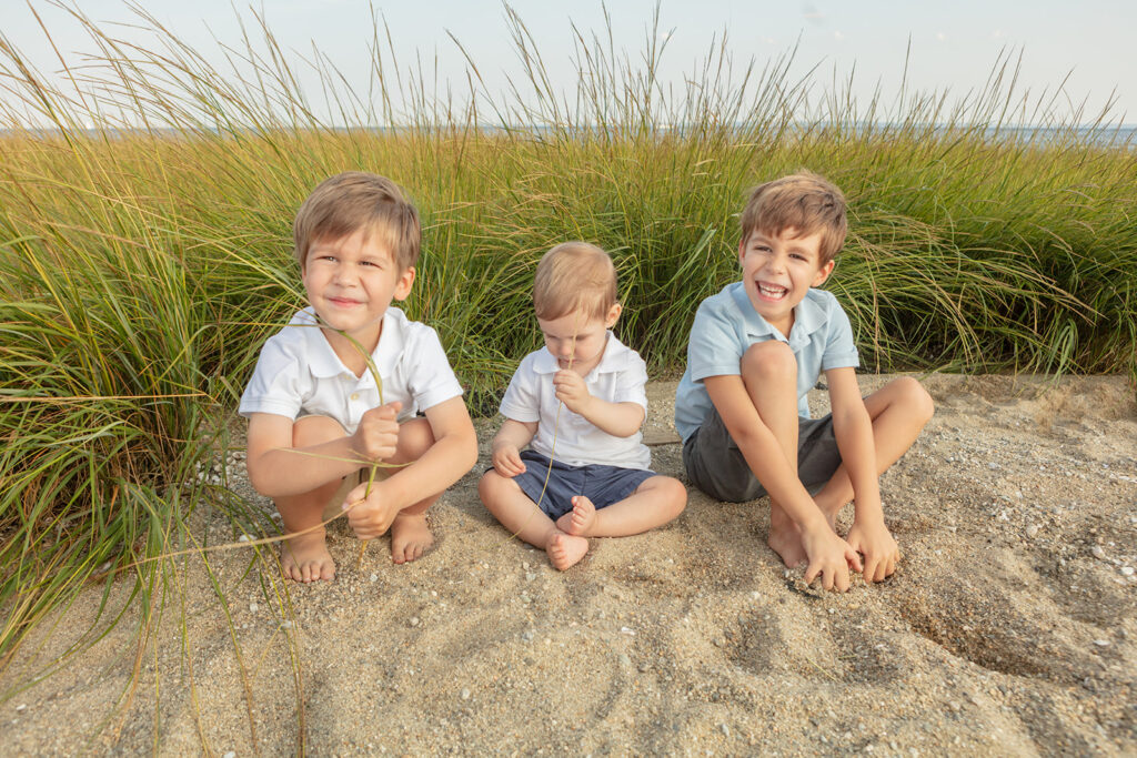 Three young boys with sandy hair sit in the sand at Greenwich Beach. Two of the boys have dune grass in their hands, while the other plays with the sand at his feet. Behind them are dune grasses and the waterline in the distance.