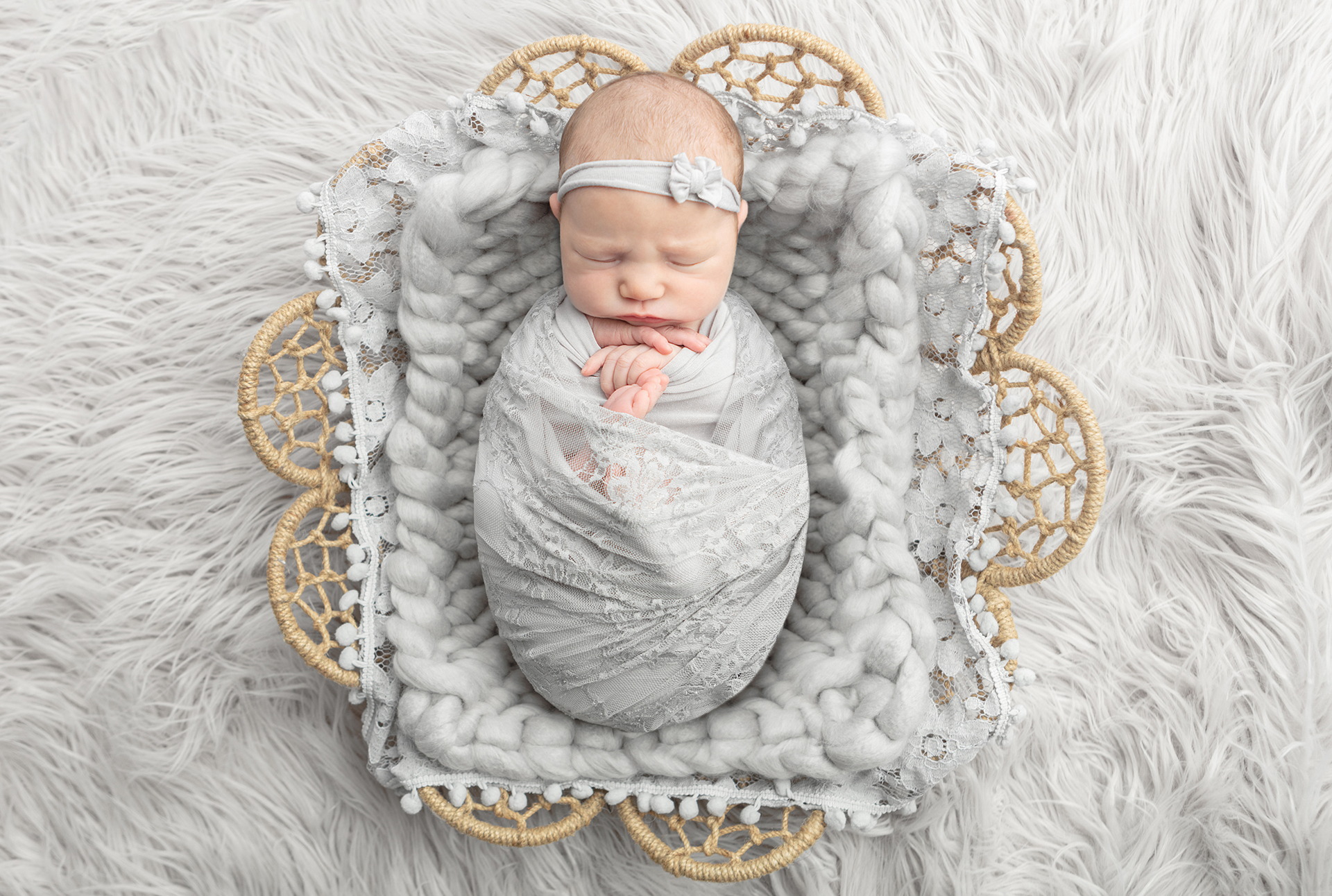 A newborn baby girl is beautifully swaddled in gray lace. She lies on a chunky silvery gray blanket, layered in a flower shaped basket, placed on a gray flokati.