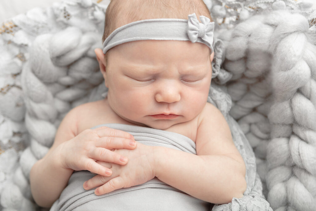 Newborn baby Olivia rests peacefully with her hands folded across her chest. She is lightly swaddled in gray and wears a matching, stretchy gray headband with a bow.