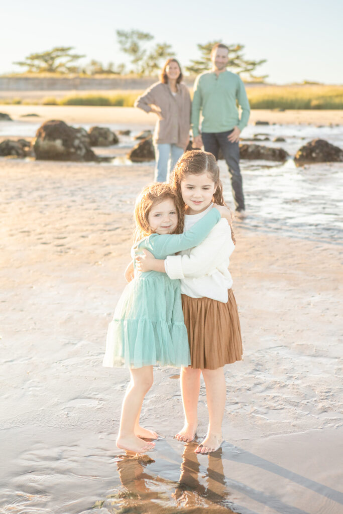 Two young sisters give each other hugs as they stand barefoot in the sand at Greenwich Beach. Their parents are out of focus in the background behind them, holding hands and smiling.