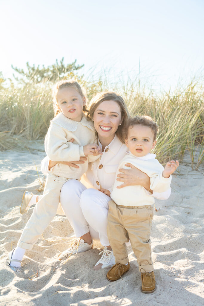 A Connecticut mother and her two young children are photographed in classically styled creams and whites, at Greenwich Beach, with sea grasses and the setting sun behind them.