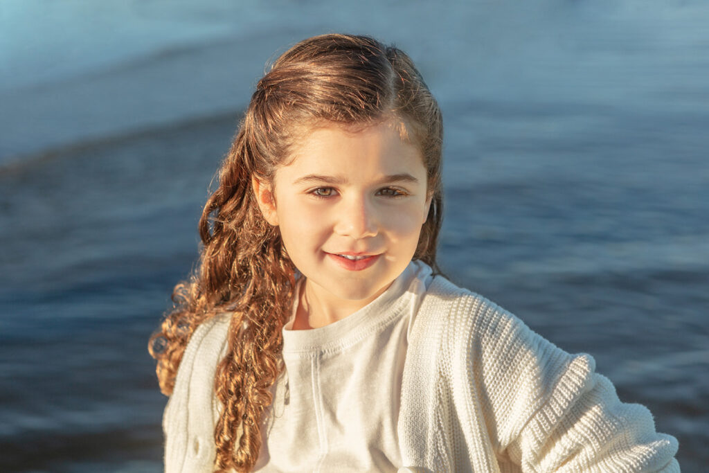 Beautiful coastal portrait of a young girl with very curly, auburn hair cascading down her shoulders. The sunlight reflects off her face, eyes, and even eyelashes, and the dark blue ocean is behind her. 