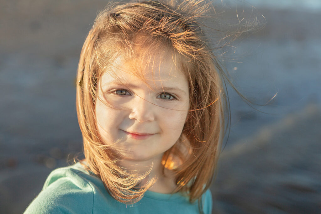 A little girl with strawberry blonde hair smirks at the camera as the wind blows her wispy hair in front of her face. The ocean glows gray-blue behind her.