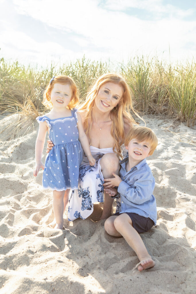 A smiling, fair mom with long, strawberry blonde hair, tilts her head and smiles as she kneels in the sand with her two young children. The trio wear coordinating blue and white outfits.