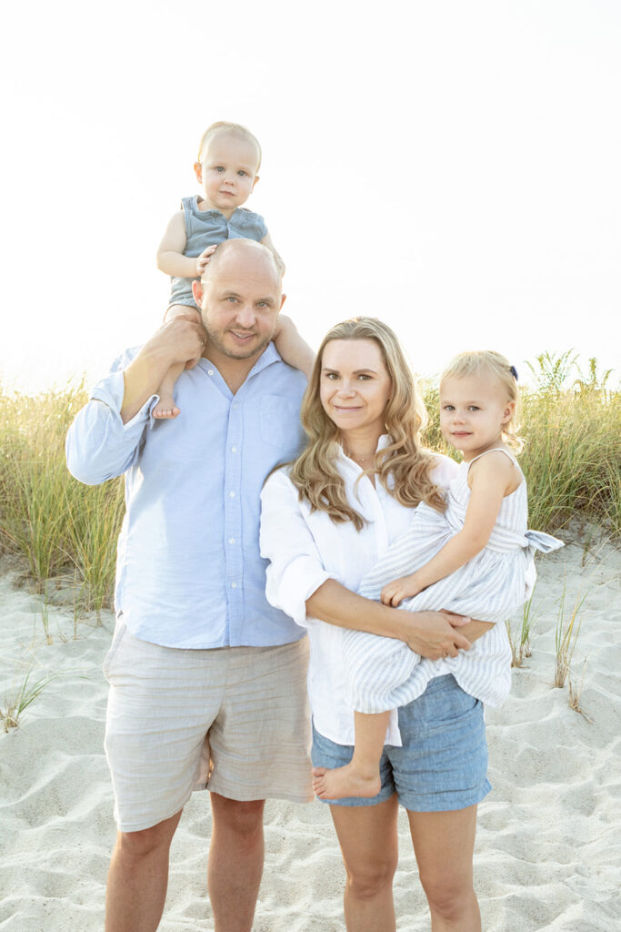 A family of four is photographed at Greenwich Beach for a classic summer family photo session. The golden sun sets behind them and the family are framed by wild beach grasses in the background.